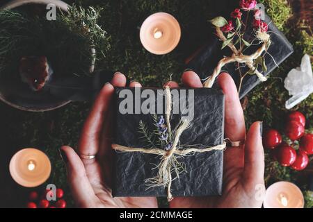 Yule Wintersonnenwende (Weihnachten) thematisierte flache Lay von weiblichen Händen Hält eine schwarze Geschenkbox in Garn und Blumen verpackt In ihren Händen Stockfoto