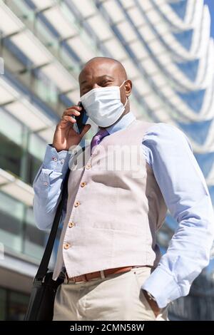 Unternehmungslustiger schwarzer Mann in eleganter Kleidung und medizinischer Maske spricht am Telefon neben einem Bürogebäude. Business und neues normales Konzept. Stockfoto