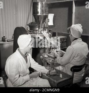 1950s, historisch, eine Arbeiterin, die mit einer Maschine mit einem Stahltrichter sitzt, kleine Glasflaschen mit medizinischen Pillen oder Tabletten füllt, während eine Kollegin die Watte hält, um an die Spitze der Pillen in der Flasche zu gehen, Brighton, England, Großbritannien. Stockfoto