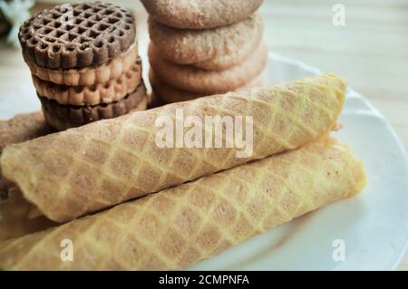 Waffelrollen und Kekse auf einem weißen Teller. Waffeln davor. Komposition auf einem hölzernen Hintergrund. Sichtbare Waffelstruktur Stockfoto