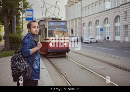An der Haltestelle des öffentlichen Nahverkehrs wartet ein Mann auf eine Straßenbahn. Ein junger Mann steht in der Nähe der Eisenbahnstrecke auf dem Hintergrund einer fahrenden Straßenbahn. Stockfoto