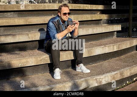 Der junge Mann mit Smartphone in den Händen sitzt auf der Beton-Treppe und sendet mit seinem Telefon Nachrichten. Stockfoto