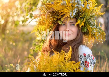 Junge schöne Frau in einem Kranz von Wildblumen und besticktes Hemd im Freien an einem sonnigen Tag. Nahaufnahme im Hochformat Stockfoto