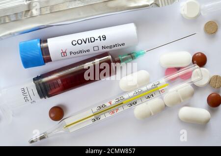 Blutproben in einer Einwegspritze und einem Reagenzglas mit einem positiven Testergebnis für COVID-19, ein medizinisches Quecksilberglas-Thermometer und verschiedene Tabletten Stockfoto