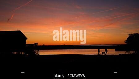 Sonnenuntergang über Assateague Island über Sümpfen, Salzwasserbucht mit Silhouette des Gebäudes, ein Paar und Hund in der Nähe des Wasserrandes. Stockfoto