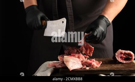 Koch in schwarzen Latexhandschuhen hält ein großes Messer und Zerschneidet rohes Kaninchenfleisch auf einem braunen Holz in Stücke Stockfoto