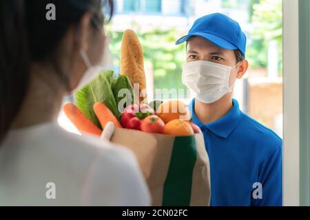Asiatische Lieferung Mann trägt Gesichtsmaske und Handschuh mit Lebensmitteln Beutel mit Lebensmitteln, Obst, Gemüse geben Frau Kunden vor dem Haus während der Zeit o Stockfoto