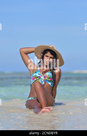 Hispanische Frau, die an einem Strand sitzt und einen schönen Bikini trägt Und Strohhut Blick auf die Kamera in Los Roques Venezuela Stockfoto