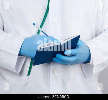 Arzt in einem weißen Mantel und blauen Handschuhen hält ein Notizbuch aus blauem Papier für Notizen Stockfoto