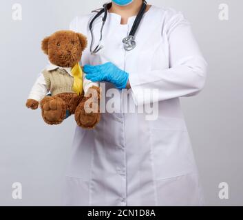 Kinderarzt in weißem Mantel, hält blaue Latexhandschuhe einen braunen Teddybären mit einem gelben Band auf einer Schwe Stockfoto