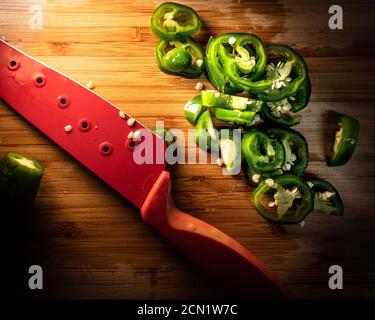 Moody kontrastreiche Aufnahme von Jalapeno Scheiben auf Holzschnitt Brett neben rotem Messer Stockfoto