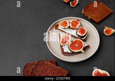 Dreieckige Sandwiches mit Feigen, Frischkäse auf schwarzem Brot mit Samen auf flachem Porzellanteller, schwarzer Tisch mit Zutaten, Draufsicht, selektiver FOC Stockfoto