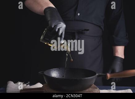 Koch in schwarzen Latexhandschuhen gießt Olivenöl aus einem Transparente Flasche in einen schwarzen Gusseisen Frittieren p Stockfoto