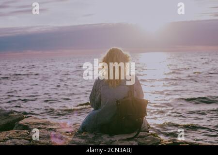 Rückansicht der blonden Frau, die am Meer sitzt Sonnenuntergang Stockfoto