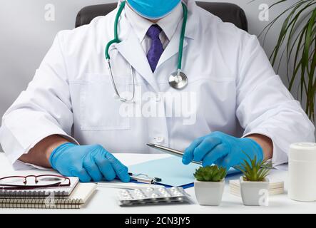 Der Arzt in weißer Uniform sitzt an einem weißen Tisch und schreibt in einem Notizbuch aus Papier, das sterile Glov trägt Stockfoto