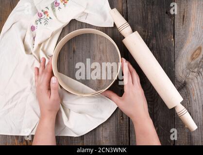 Weibliche Hände halten ein rundes Holzsieb für Mehl, neben einem hölzernen Nudelholz Stockfoto