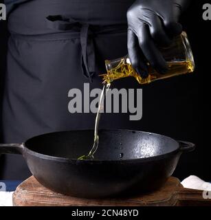 Koch in schwarzen Latexhandschuhen gießt Olivenöl aus einem Transparente Flasche in einen schwarzen Gusseisen Frittieren p Stockfoto