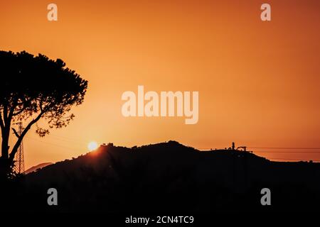 Berge, Zedernkronenbaum und Turm mit Hochspannungsdrähten Silhouetten bei schönem Sonnenuntergang mit orangefarbenem Himmel und Sonnenuntergang über dem Berg. Kontrast Stockfoto