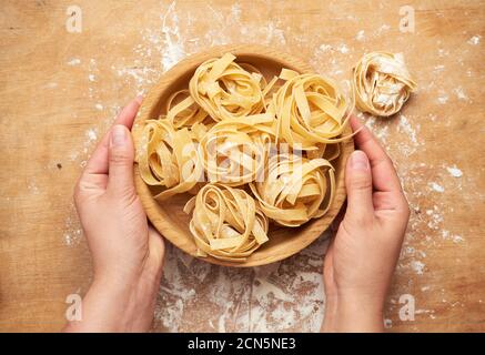 Weibliche Hände halten einen Holzteller mit roher Fettuccine Pasta Über einem Holztisch Stockfoto