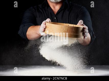 Koch ein Mann in schwarzer Uniform hält eine Runde Holzsieb in den Händen und siebt Weizenmehl Stockfoto