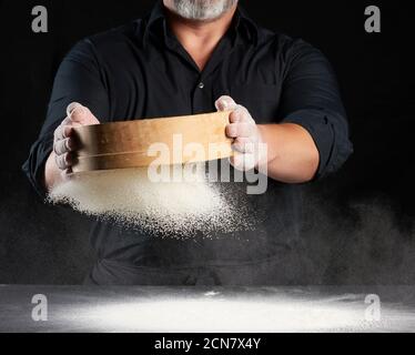 Koch ein Mann in schwarzer Uniform hält eine Runde Holzsieb in den Händen und siebt Weizenmehl Stockfoto