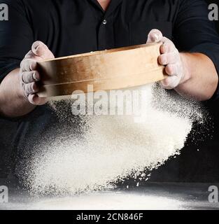 Koch ein Mann in schwarzer Uniform hält eine Runde Holzsieb in den Händen und siebt Weizenmehl Ein Stockfoto