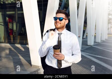 Geschäftsmann zu Fuß am sonnigen Tag mit Jacke auf der Schulter. Geschäftsmann lächelt mit Sonnenbrillen und Coffee to Go Tasse. Stockfoto