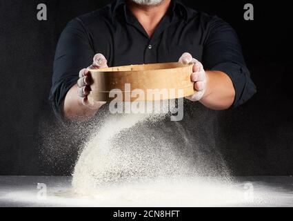 Koch ein Mann in schwarzer Uniform hält eine Runde Holzsieb in den Händen und siebt Weizenmehl Ein Stockfoto