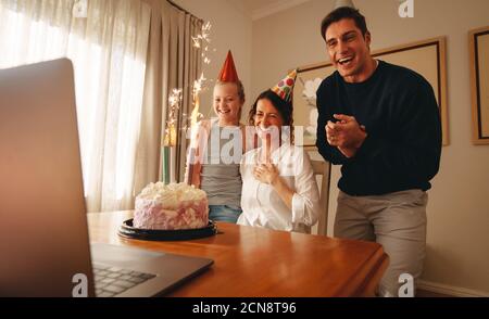 Frau feiert Geburtstag zu Hause mit Familie und Freunden auf Videoanruf. Laptop und Geburtstagskuchen mit Kerzen auf dem Tisch mit Familie trägt Party Hut Stockfoto