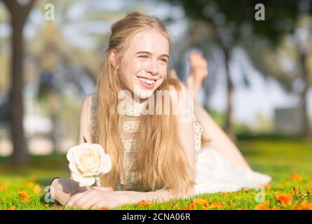Happy hübsche junge Teenager Mädchen auf dem grünen Gras liegen, während eine weiße Rose im Park während eines sonnigen Tages halten. Stockfoto