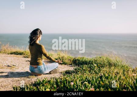 Ruhige Frau meditiert auf sonnigen Klippe mit Blick auf das Meer Stockfoto