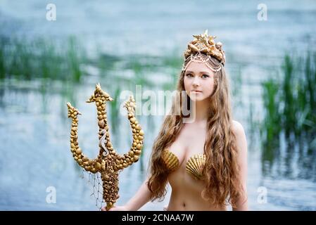 Fantasy Frau echte Meerjungfrau mit Dreizack Mythos Göttin des Meeres mit goldenen Schwanz sitzt in Sonnenuntergang auf Felsen.. Gold Haar Krone Muscheln Perlen Schmuck. Merma Stockfoto