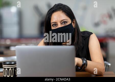 Porträt einer Inderin mit Covid 19 Schutzmaske und Arbeit an ihrem Laptop, sitzen in einer Bürokantine, Café. Stockfoto