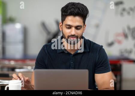 Porträt eines jungen hübschen indischen Mannes, der an seinem Laptop arbeitet, in einer Bürokantine sitzt, in einem Café, in einer ungezwungenen Arbeitsumgebung. Stockfoto