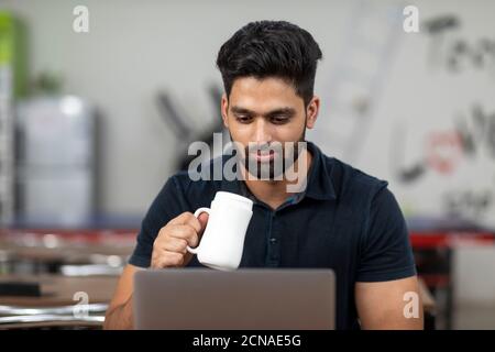 Porträt eines jungen, gutaussehenden indischen Mannes, der eine Kaffeetasse hält und an seinem Laptop arbeitet, in einer Bürokantine sitzt, in einem Café, in einer legeren Arbeit. Stockfoto