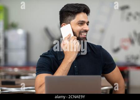 Porträt eines jungen hübschen indischen Mannes, der am Telefon spricht und am Laptop arbeitet, in einer Bürokantine sitzt, in einem Café, in einer legeren Arbeit. Stockfoto