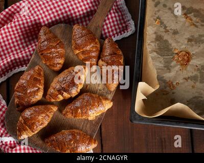 Gebackene knusprige Croissants auf einem braunen Holzbrett, neben einem Backblech mit Pergamentpapier Stockfoto