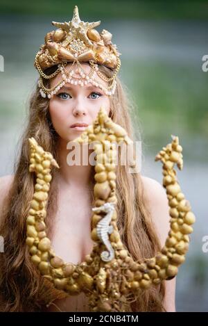 Fantasy Frau echte Meerjungfrau mit Dreizack Mythos Göttin des Meeres mit goldenen Schwanz sitzt in Sonnenuntergang auf Felsen.. Gold Haar Krone Muscheln Perlen Schmuck. Merma Stockfoto