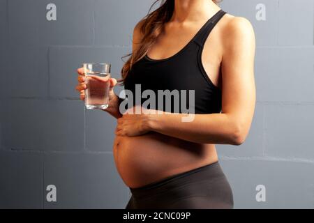 Schwangere Frau im Sport-Workout-Outfit mit einem Glas Wasser. Eine Nahaufnahme des Bauches einer schwangeren Frau. Gesunde Ernährung während der Schwangerschaft. Stockfoto