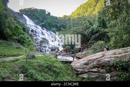 DOI INTHANON NATIONAL PARK, THAILAND - Sep 02, 2019: Eine Frau sitzt auf einem Felsen gegenüber dem hoch aufragenden Mae Ya Wasserfall in Thailand. Stockfoto
