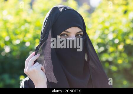 Eine Migrantin aus dem Osten in schwarzer Kleidung. Eine Migrantin aus dem Osten in schwarzer Kleidung steht vor leuchtend grünen Bäumen. Stockfoto