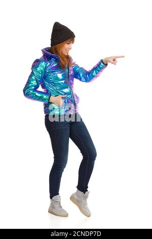 Die junge Frau in lebhafter und glänzender Daunenjacke, Mütze, Jeans und Sneakers schaut weg, zeigt und berührt etwas mit dem Zeigefinger. Seitenansicht. F Stockfoto