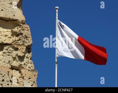 Flagge von Malta, die gegen einen blauen Himmel durch Sandsteinmauer in Valletta, Malta fliegt Stockfoto