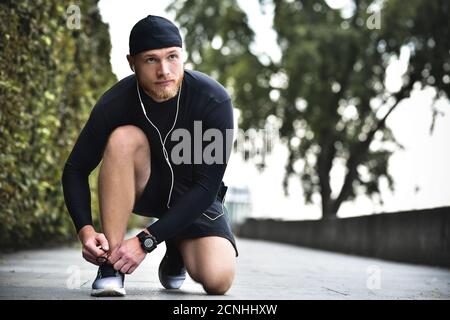 Ernsthafter bärtiger kaukasischer junger Sportler, der im Stadtpark Schnürsenkel auf seine Sneakers bindet. Stockfoto