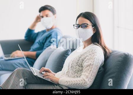 Nette hübsche asiatische Frau trägt medizinische Schutzmaske auf Gesicht mit Laptop Notebook-Computer zu Hause im Wohnzimmer mit kaukasischen Mann arbeiten. Idee Stockfoto