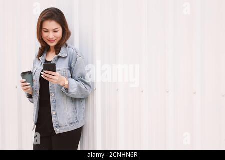 Hübsche asiatische Frau in Jeans stehend hält Kaffeetasse und mit Smartphone mit fröhlich fröhliche Art und Weise. Idee für die Arbeit am Morgen mit Pow gehen Stockfoto