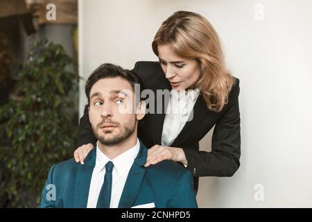 Weibliche Chefin verführt Angestellte. Das junge Mädchen hält an den Schultern eines Kollegen, der am Tisch sitzt. Er sieht sie überrascht an. Hohe Qualität Stockfoto