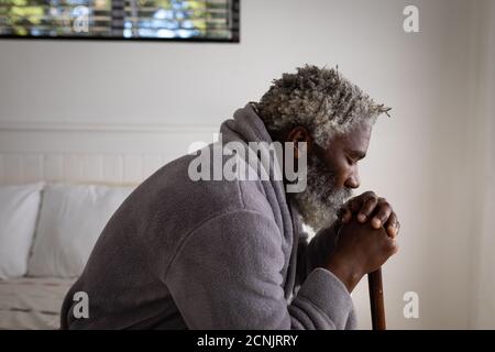 Afroamerikanischer älterer Mann, der auf einem Bett in einem Schlafzimmer sitzt, seinen Kopf auf einem Stock ruht, soziale Distanzierung und Selbstisolierung in Quarantänesperre Stockfoto