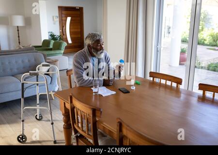 Afroamerikanischer älterer Mann sitzt an einem Tisch im Wohnzimmer, hält zwei medizinische Pillen und ein Glas Wasser in den Händen, soziale Distanzierung und se Stockfoto
