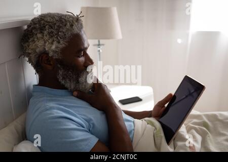 Afroamerikanischer Senioren, der auf einem Bett in einem Schlafzimmer liegt, mit einem digitalen Tablet, sich das Kinn reibt, soziale Distanzierung und Selbstisolation in Quarantäne Stockfoto